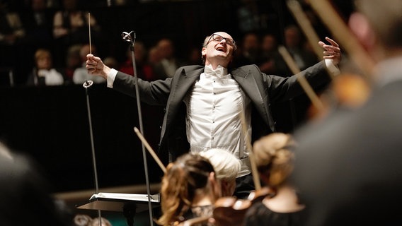 Der Dirigent Andrew Manze steht mit ausgebreiteten Armen vor dem Orchester © NDR Foto: Micha Neugebauer