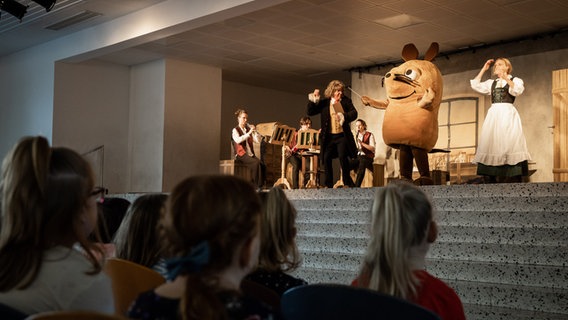 Die Maus steht auf der Bühne einer Grundschule und dirigiert. © NDR Foto: Helge Krückeberg