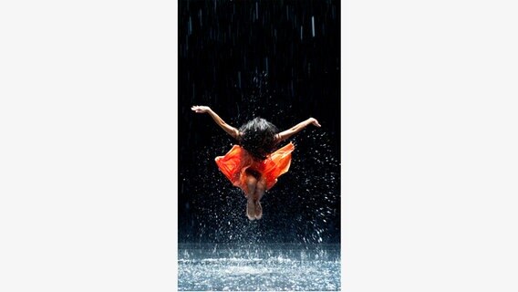 Eine Frau im orangenen Kleid springt hoch und breitet die Arme aus - Szene aus dem Film "Pina" © NDR/Wim Wenders Stiftung 