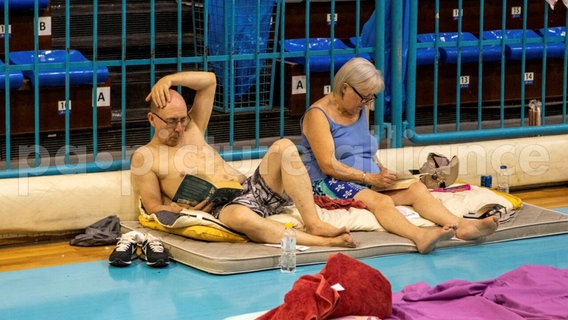 Zwei Urlauber liegen auf einer Matratze in einer Turnhalle. © picture alliance/dpa/InTime News/AP | Lefteris Damianidis 