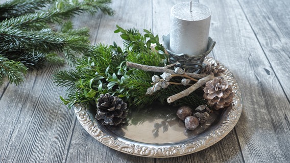 Eine silberne Kerze steht auf silbernen Tablett mit Tannengrün und Zapfen dekoriert. © NDR Foto: Anja Deuble