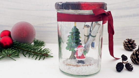 In einem Marmeladenglas steht ein kleiner Weihnachtsmann neben einer Tanne, dahinter sind gerollte Geldscheine zu sehen. © NDR Foto: Anja Deuble