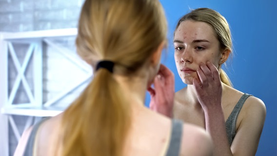 Eine junge Frau mit Akne im Gesicht betrachtet sich kritisch in einem Spiegel. © Colourbox Foto: Ievgen Chabanov