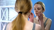Eine junge Frau mit Akne im Gesicht betrachtet sich kritisch in einem Spiegel. © Colourbox Foto: Ievgen Chabanov
