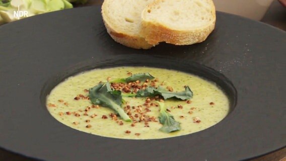 Suppe aus Brokkoli-Röschen angerichtet auf einem schwarzen Teller mit Baguette.  