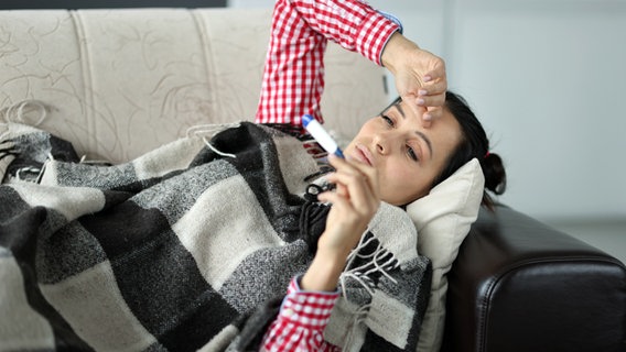 Eine Frau liegt krank mit Wolldecke auf einem Sofa und schaut auf ein Fieberthermometer. © Colourbox 
