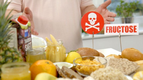 Ein Totenkopf-Schild mit der Aufschrift "Fruktose" steht neben Brötchen und anderen Nahrungsmitteln. © NDR 