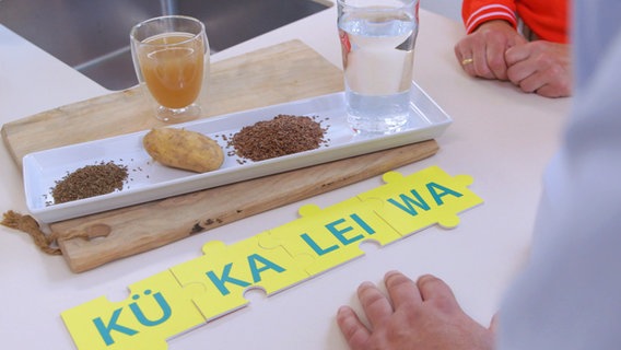 Silbenpuzzle "Kükaleiwa" liegt auf dem Tisch, dahinter Kümmelsamen, Kartoffel, Leinsamen und Wasser. © NDR 
