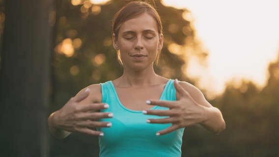 Eine Frau steht in der Dämmerung in der Natur und führt eine Qi Gong Übung aus. Die Hände sind vor ihrer Brust angehoben. © IMAGO / Depositphotos 