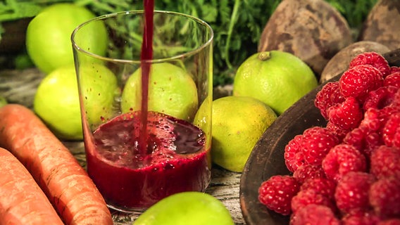Ein selbstgemachter Rote-Bete-Shot im Glas steht neben Himbeeren, Limetten und Karotten.  