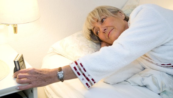 Eine ältere Frau liegt im Bett und schaut besorgt auf ihren Wecker. © picture alliance Foto: chromorange