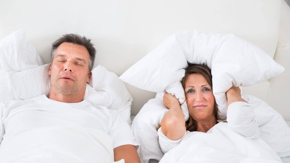 Eine Frau und ein Mann liegen im Bett, der Mann schnarcht, die Frau hält sich ein Kissen vor die Ohren. © picture alliance / dpa Themendienst | Christin Klose 
