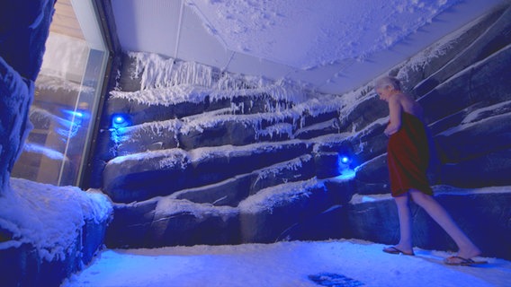 Frau, nur mit Handtuch und Badelatschen bekleidet, geht durch einen blau beleuchteten Raum mit Eiszapfen. © NDR Foto: Oliver Zydek