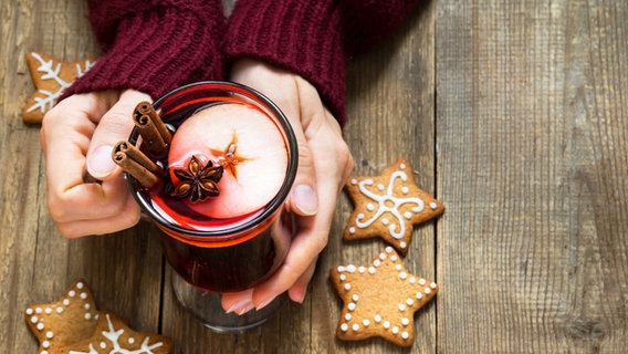 Eine Frau sitzt am Tisch und hält ein Glas Glühwein in den Händen, daneben liegen ein paar Weihnachtskekse. © Colourbox Foto: -