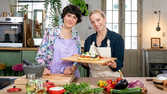 Zora Klipp und Theresa Knipschild stehen mit einem Brett Blätterteig-Pizza in einer Küche. © NDR Foto: Claudia Timmann