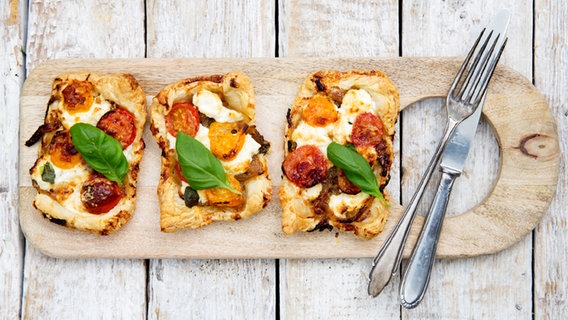 Drei kleine Tartes mit Tomaten, Ziegenkäse und Basilikum liegen auf einem Holzbrett. © NDR Foto: Claudia Timmann