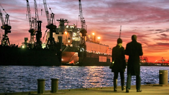 Ein Paar steht an den Landungsbrücken im Hamburger Hafen und blickt auf die Elbe mit dem Schwimmdock und den stimmungsvollen Abendhimmel. © dpa Foto: Bodo Marks