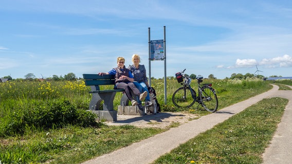 Zwei fröhliche Damen sitzen auf einer sogenannten "Baumelbank" am Hebbelrundwanderweg bei Norddeich © NDR Foto: Christine Raczka