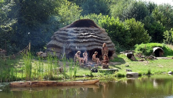 Mitarbeiter sitzen in steinzeitlicher Kleidung vor einer rekonstuierten Hütte auf dem Gelände des Steinzeitparks Dithmarschen. © Steinzeitpark Dithmarschen 