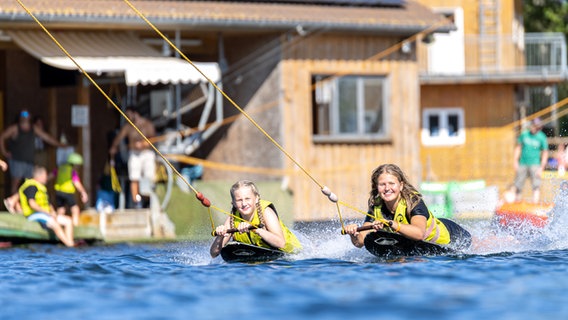 Zwei Mädchen liegen auf Wakeboards und lassen sich von einem Wasserskilift ziehen auf. © Alfsee Wasserski GmbH Foto: Christian Senft