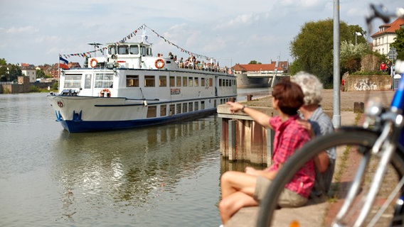 Touristen sitzen am Ufer der Weser in Hameln und schauen auf ein vorbeifahrendes Weserschiff. © Weserbergland Tourismus e.V. 