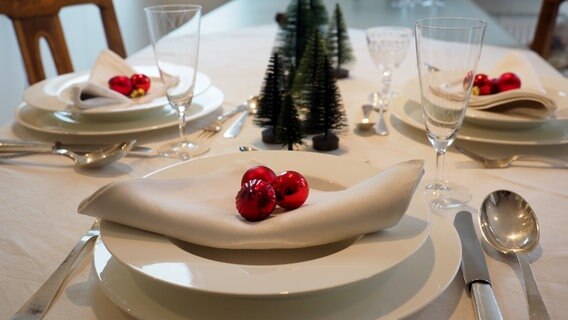 Ein festlich gedeckter Tisch mit weißem Geschirr, weißen Stoffservietten, roten Christbaumkugeln und grünen Tannenbäumchen. © NDR Foto: Anja Deuble
