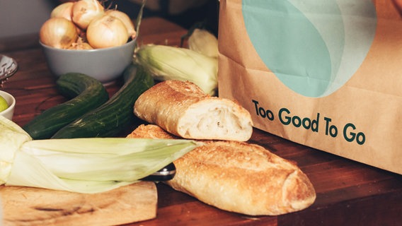 Gemüse und Baguette liegen in einer Küche, daneben eine Papiertüte mit dem Logo der Organisation "Too Good To Go". © Too Good To Go 
