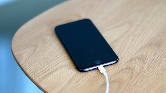 Ein schwarzes Smartphone auf einem Holztisch mit eingestecktem Ladekabel © NDR Foto: Elke Janning