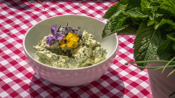 Kräuterbutter in einem Schälchen, mit Blüten verziert. Daneben sind einige Kräuter im Anschnitt zu sehen. © NDR 