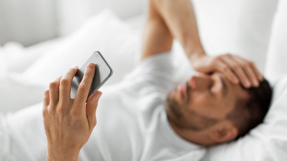 Ein im Bett liegender Mann hält sich mit einer Hand den Kopf, in der anderen ein Smartphone © colourbox 