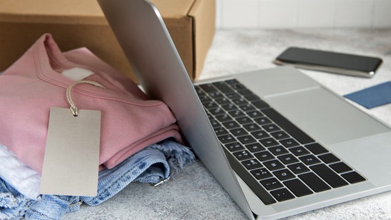 Ein Laptop, daneben Modeartikel und ein Versandpaket auf hellgrauer Arbeitsplatte © colourbox 