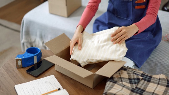 Eine Frau legt ein Kleidungsstück in einen geöffneten Pappkarton, daneben Klebeband und Handy © colourbox 
