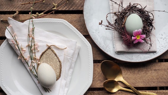 Zwei weiße Teller mit Leinenservietten, Eiern und einem kleinen Kranz stehen auf einem alten Holztisch. © NDR Foto: Anja Deuble