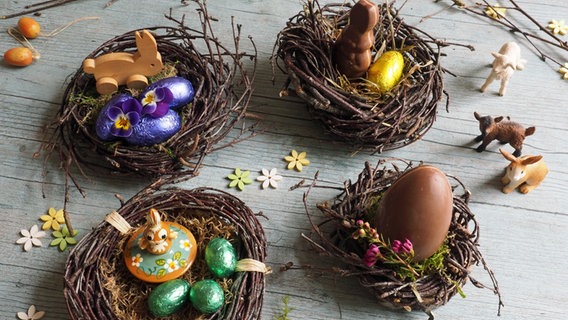 Vier kleine Vogelnester mit Schokoladeneiern, kleinen Spielzeugen und bunten Blüten liegen auf einem Tisch. © NDR Foto: Anja Deuble