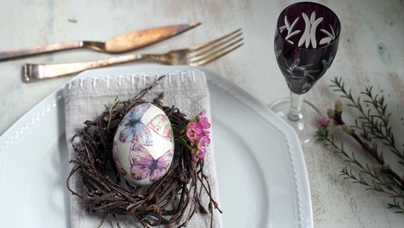 Ein mit Schmetterlingen gestaltetes Ei liegt in einem Nest aus Birkenreisig auf einem Teller. © NDR Foto: Anja Deuble