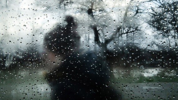 Eine Person mit Pudelmütze geht hinter einer beschlagenen und mit Regentropfen gesprenkelten Fensterscheibe entlang. © picture alliance/Bernd Thissen/dpa Foto: Bernd Thissen