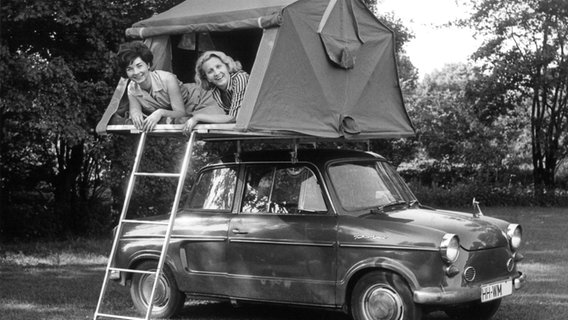 Zwei Frauen liegen auf einem historischen schwarz-weiß-Foto in einem Zelt auf einem Autodach und lachen in die Kamera. © picture alliance Foto: Georg Spring