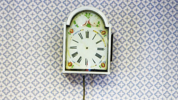Eine Uhr ohne Zeiger hängt an einer Wand. Die Tapete ist blau weiß gemustert und wirkt alt. © Julia Schulz 