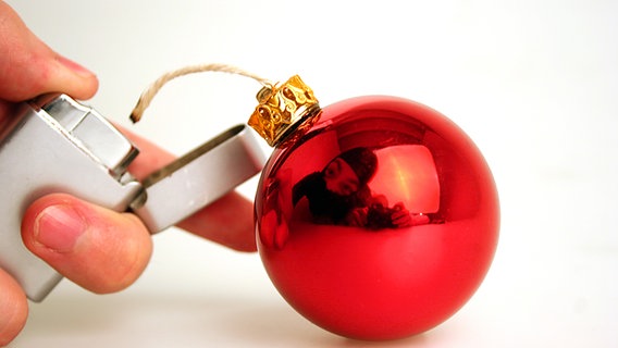 Eine rote Weihnachtskugel aus der ein Docht hängt. Eine Hand hält ein Feuerzeug dass den Docht anzündet. in der Spiegelung der Kugel ist eine teilweise maskierte Person zu sehen. © photocase.de Foto: PERFFZERFF
