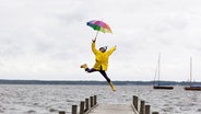 Eine Frau in Regenjacke und mit Regenschirm springt auf einem Steg am Wasser © NDR Foto: Westend61