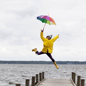 Eine Frau in Regenjacke und mit Regenschirm springt auf einem Steg am Wasser © NDR Foto: Westend61