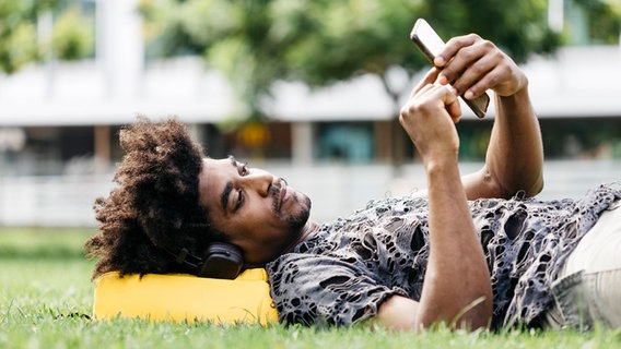 Ein Mann liegt auf einer Wiese, trägt Kopfhörer und hört mit seinem Smartphone Musik. © imago images Foto: Westend61