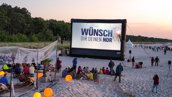 Menschen am Strand schauen am Abend auf eine Videoleinwand mit dem Logo der Aktion "Wünsch Dir Deinen NDR".  (Montage). © NDR Foto: Anja Deuble / NDR